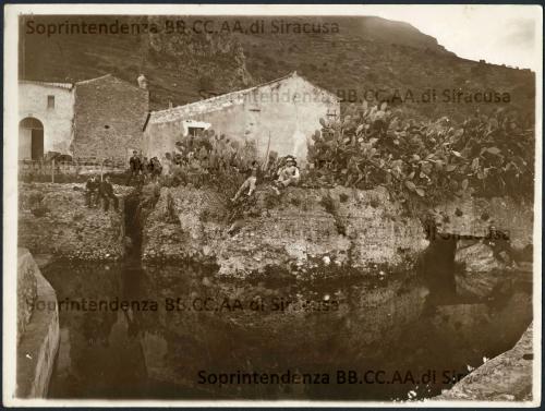 Salvo, Michele (1876 - 1963), Italia, Sicilia, Termini Imerese (PA), località Brucato - “Sorgenti di Brucato dove i romani cominciarono a costruire l’acquedotto Cornelio che portava le acque a Termini”, la fotografia è animata da tre gruppi di uomini e adolescenti e sulla destra un uomo in disparte. L’intento del fotografo è quello di documentare le sorgenti del Brucato e di curare l’aspetto artistico della composizione sottolineando come questa si rifletta nell’acqua della sorgente. Sul verso della fotografia sono indicati: il proprietario della sorgente Comune di Termini Imerese, le informazioni geografiche e l’autore dello scatto fotografico Salvo Michele che durante la sua attività documenta non solo aspetti del suo paese ma anche la Targa Florio, nel 1905 i Musei Vaticani e i disastri provocati a Messina dal terremoto del 1908. Il suo Archivio Fotografico è stato recuperato e conservato dopo la sua morte, avvenuta il 31 maggio 1963, dalla famiglia Ciofalo, collocazione Fondo Giuseppe Cultrera C6 ID 1474 - Nel Fondo sono presenti altre immagini dell'autore riguardanti Termini Imerese: “ruderi della Curia Romana nella Villa Comunale Nicolò Palmeri”, “ruderi dell’Anfiteatro” e “dintorni”, XIX - XX secolo, Materia: carta, Tecnica: stampa su carta alla gelatina al bromuro d'argento virata seppia, mm 179 x 239 mm., CC BY-NC-ND