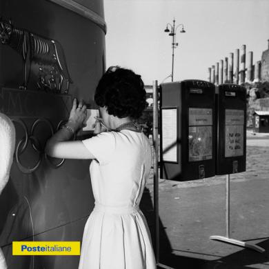 1960, Roma. Una donna scrive una cartolina appoggiandosi alla fiancata di un ufficio postale mobile in servizio in occasione delle XVII Olimpiadi, CC BY-NC-ND