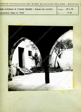 Archivio fotografico Galleria Reg. di Palazzo Bellomo, Stampa su carta fotografica in bianco e nero, in formato 18X24 cm., risalente al 1935 circa, posta su cartoncino dell’Archivio fotografico della Galleria Reg. di Palazzo Bellomo, già Museo Nazionale. Il soggetto è una veduta del cortile occidentale del palazzo dal portico dell’ingresso principale. L’autore non è indicato., Stampa in B/N da pellicola, CC BY-NC-ND