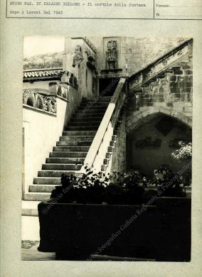 Archivio fotografico Galleria Reg. di Palazzo Bellomo, Stampa su carta fotografica in bianco e nero di formato non indicato, risalente al 1942 circa, ripresa dopo alcuni lavori di restauro del palazzo, atti ad istituire il Museo Nazionale. La stampa è posta su cartoncino dell’Archivio fotografico della Galleria Reg. di Palazzo Bellomo, già Museo Nazionale. Il soggetto è una veduta del Cortile della Fontana, ripreso dal portico al piano terra del palazzo. Si può notare la scala in stile catalano, con l’edicola e lo stemma della famiglia Bellomo sul muro, sopra la prima rampa. L’autore non è indicato., Stampa in B/N da pellicola, CC BY-NC-ND