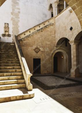 Archivio fotografico Galleria Reg. di Palazzo Bellomo, Immagine digitale a colori, anno 2024, ripresa dal portico del piano terra, con veduta sulla scala in stile catalano, nel Cortile della Fontana. Si intravede la loggetta al piano superiore, ricostruita in stile originario nel 1952. Autore: G. Gallitto., Ripresa in esterni, CC BY-NC-ND