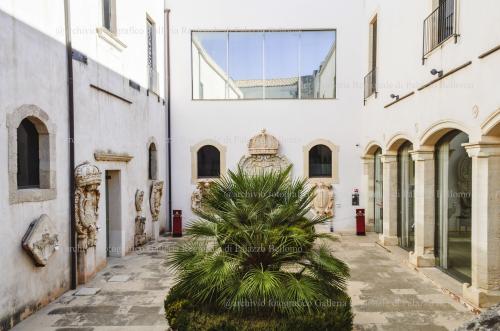 Archivio fotografico Galleria Reg. di Palazzo Bellomo, Immagine digitale a colori, anno 2024, ripresa da sopra la prima rampa della scala di Palazzo Bellomo con veduta sul Cortile degli Stemmi. Si notano i molteplici stemmi gentilizi e reali esposti lungo le due pareti del cortile, a destra il portico con le vetrate sede di esposizione della Galleria Reg. di Palazzo Bellomo. Autore: G. Gallitto., Ripresa in esterni, CC BY-NC-ND