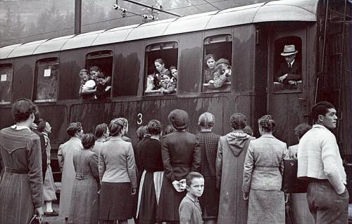 Partenza di un treno: diverse ragazze salutano dai finestrini di un vagone le persone sul marciapiede. BRESSANONE, stazione ferroviaria COLLEZIONE ROMEO SOMMAVILLA, n. 765, CC BY-SA
